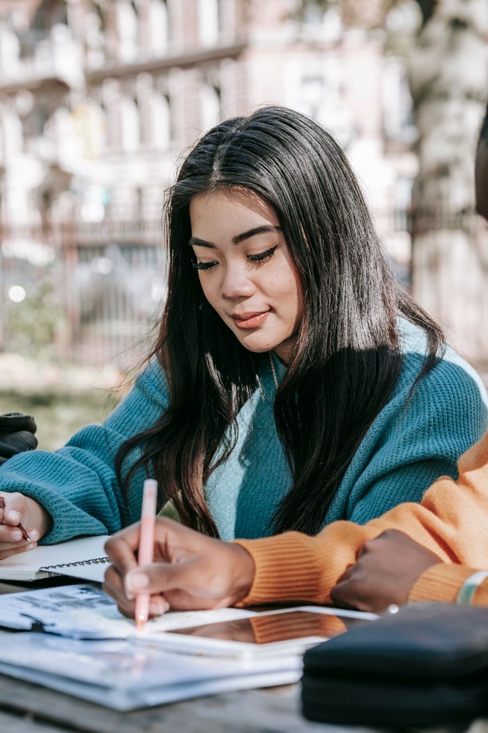 services-02 Content young multiracial female students in warm sweaters working on university project together and taking notes in copybooks while sitting at table in park