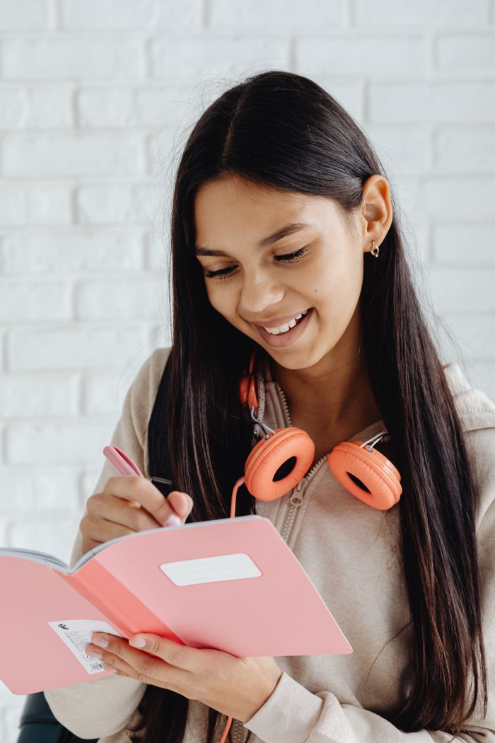 Teen girl writes in notebook wearing headphones, smiling indoors.