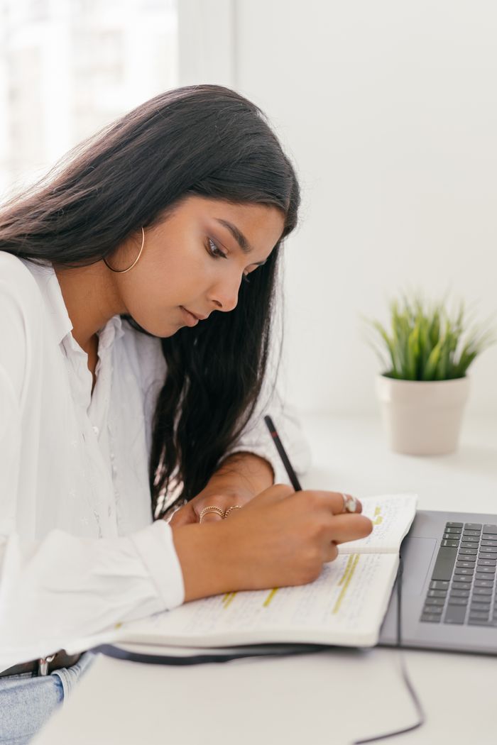 services-01 Young woman intensely writing notes in a notebook at home, engaging in online learning with a laptop.